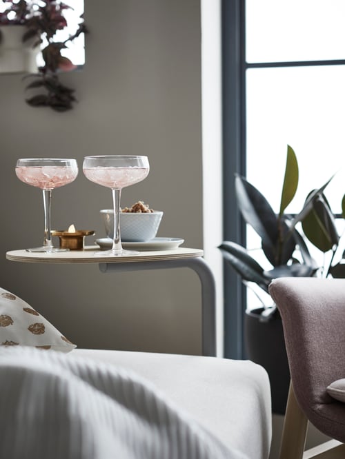 Two clear glass, STORHET champagne coupes hold pink drinks on a side table with a bowl of snacks, attached to a sofa-bed.