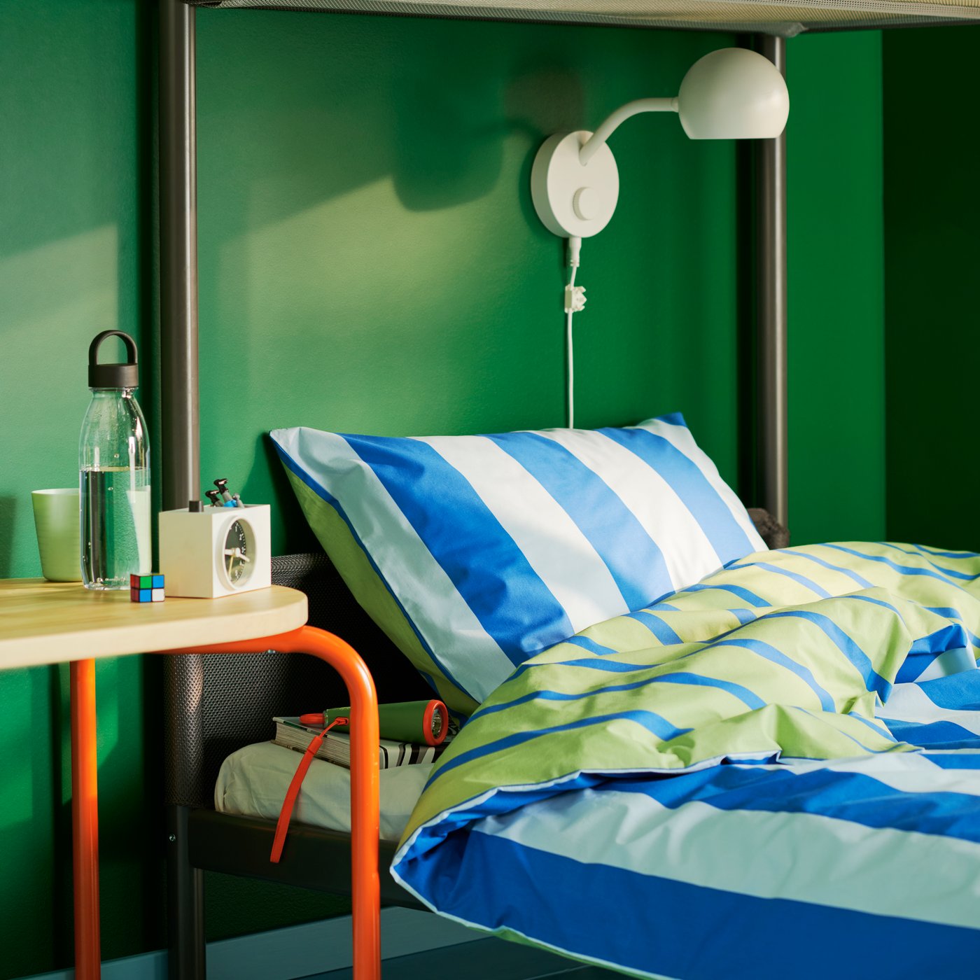 A striped BARNDRÖM duvet cover and pillow case, on a bed in a green-walled children’s room with a lamp above the bed.
