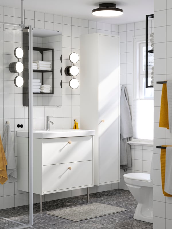 A white HAVBÄCK wash-stand and a white HAVBÄCK high cabinet are standing in a white bathroom with white and yellow towels.