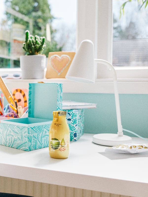 A MUNSBIT oat smoothie stands on a desk by a window along with storage boxes, pens and a white FUBBLA work lamp.