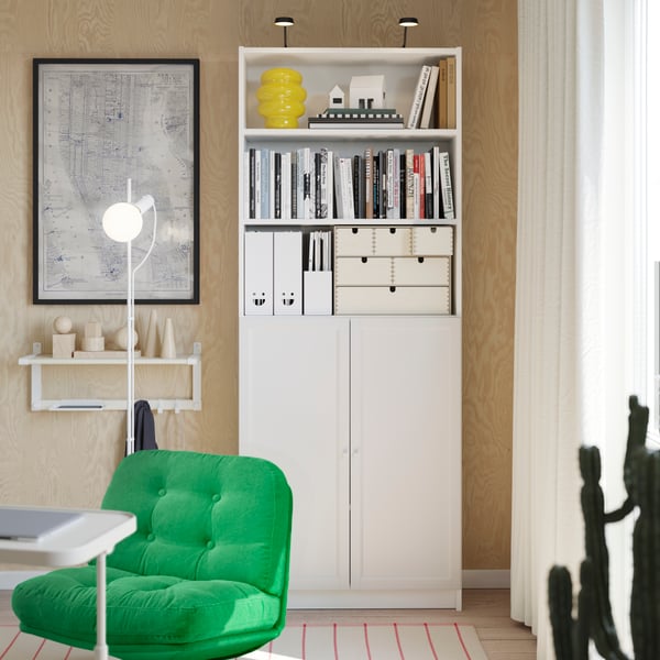 A white BILLY bookcase and a small white shelf hanging next to it, with a white floor lamp standing in the front.