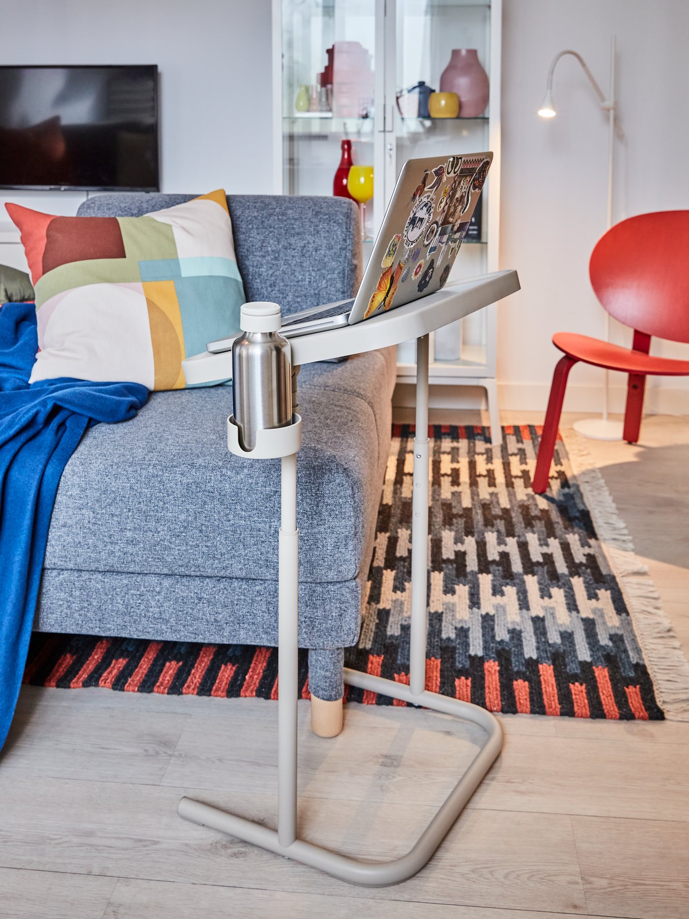 Malý přenosný stolek u šedé pohovky, na stolku notebook a termo láhev, moderní obývací pokoj s barevným polštářem, modrou dekou a ručně vyrobeným, vícebarevným kobercem RESENSTAD.