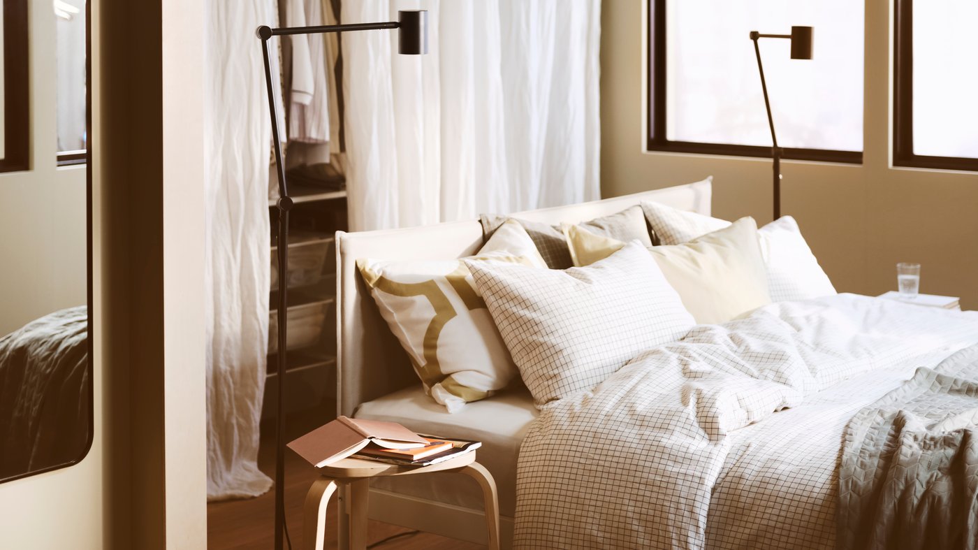 Camera da letto con struttura letto imbottita KLEPPSTAD bianco beige, due lampade da terra di colore nero, con biancheria da letto bianca.