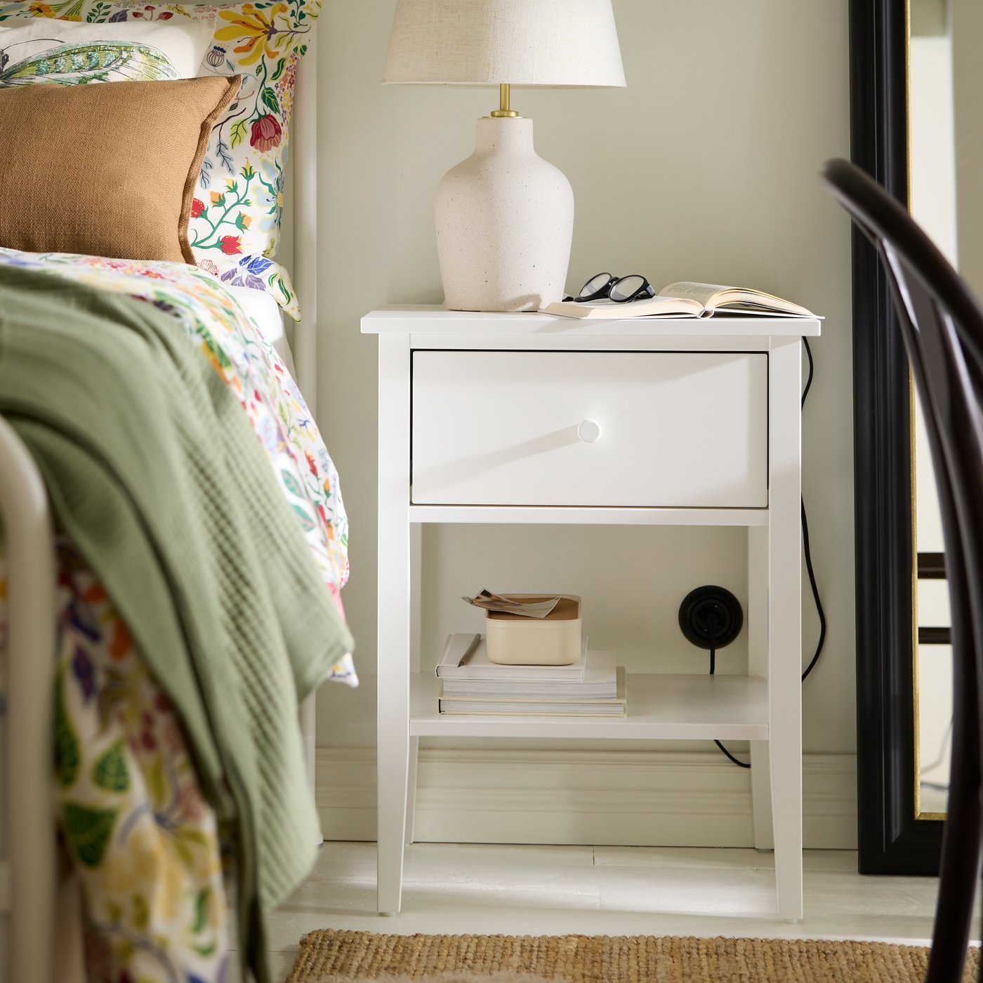 A GULLABERG bedside table in white, holding a lamp, reading glasses on a book, with a lower shelf with smaller items.