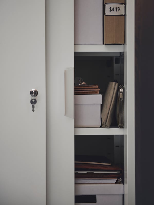 Chave pendurada na fechadura de um armário com portas deslizantes TROTTEN, em branco, parcialmente aberto, a revelar papel e caixas TJENA no interior.