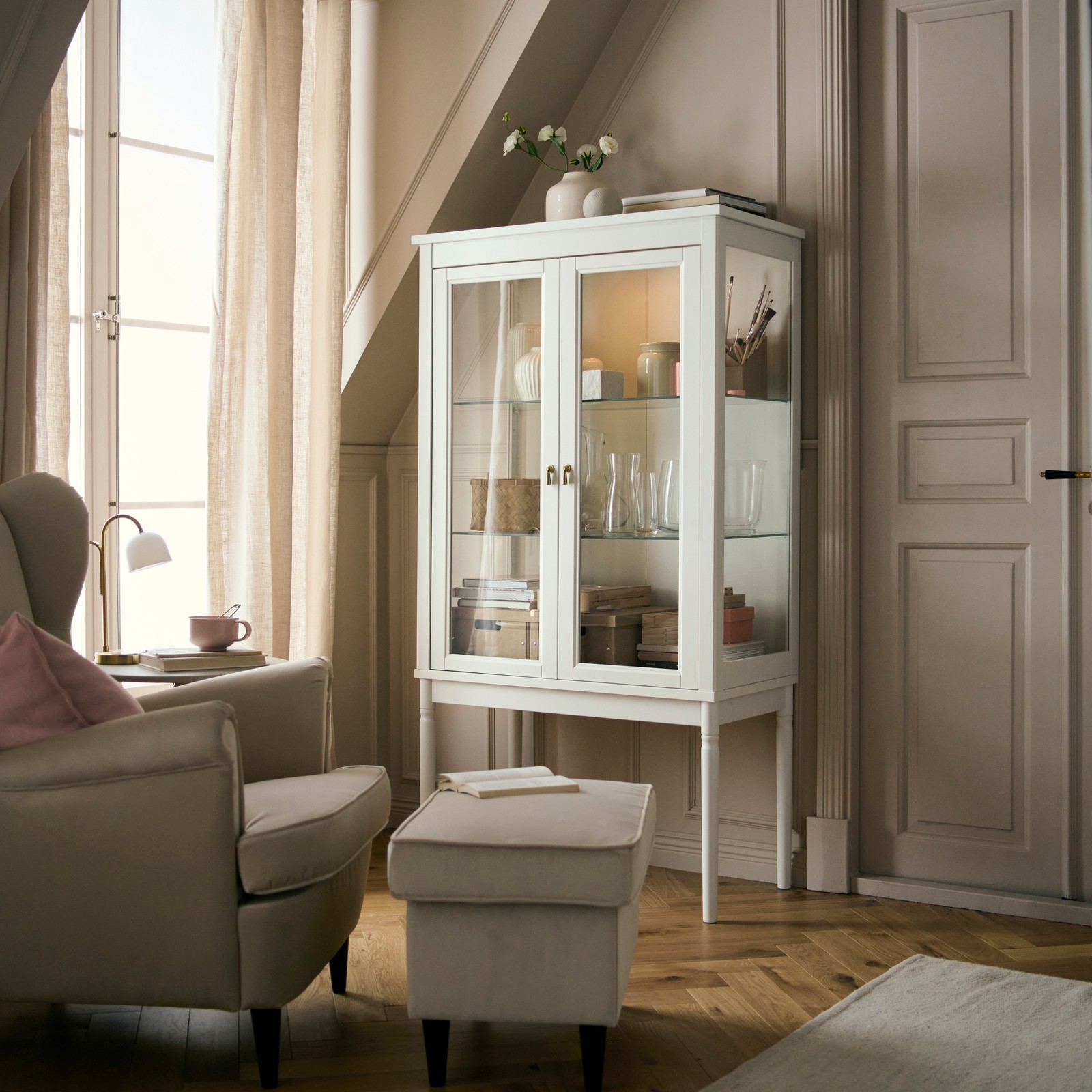 A FÄRJKARL glass-door cabinet with vases and other items on its shelves stands in a corner near a window.