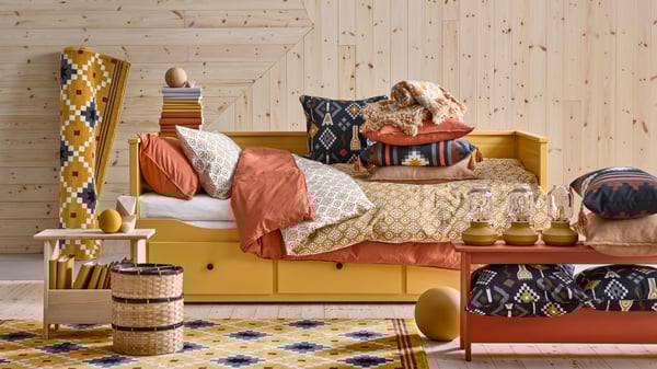 Lit banquette HEMNES jaune avec coussins, plaids et linge de lit. Il s’agit entre autres des nouveaux coussins MOSSNYCKLAR et FRUKTMOTT.