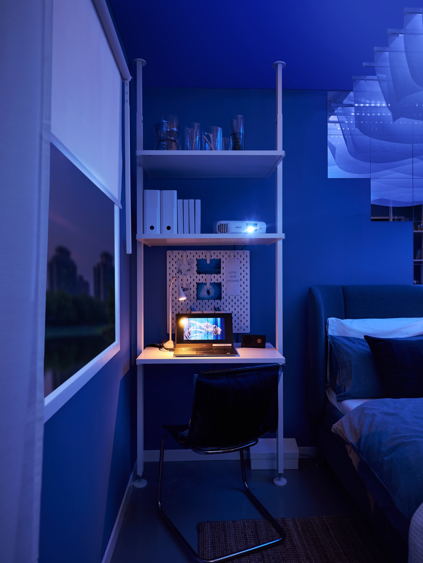 Ett sovrum i blå belysning med en säng, kuddar och filt, samt ett litet skrivbord med hyllor med en laptop på och en stol bredvid.