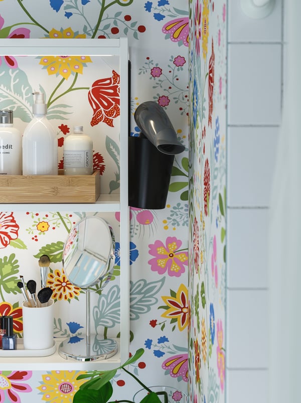 Un portaoggetti SKATTÅN con all'interno un phon appesa su una parete fantasia. Ci sono mensole appese con un vassoio TAVELÅN, uno specchio TRENSUM e altri oggetti come amalti, pennelli e altro.