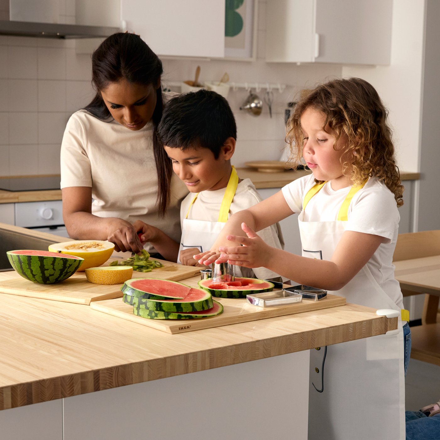 A woman and two children wearing aprons, standing by a kitchen counter cutting melon and watermelon in pieces.