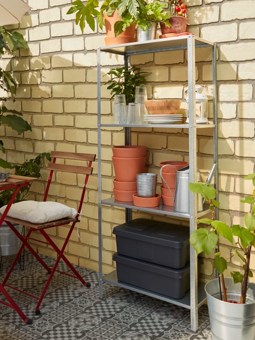 A shelving unit with four shelves holding storage boxes, plant pots, plates and glasses within easy-reach of the table.