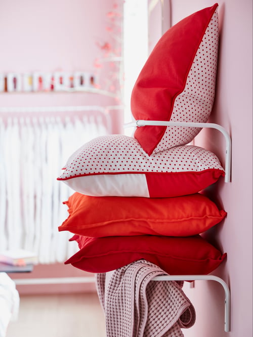 Two white MULIG clothes bars on top of each other, on a wall with cushions stacked in between and a blanket draped over one.