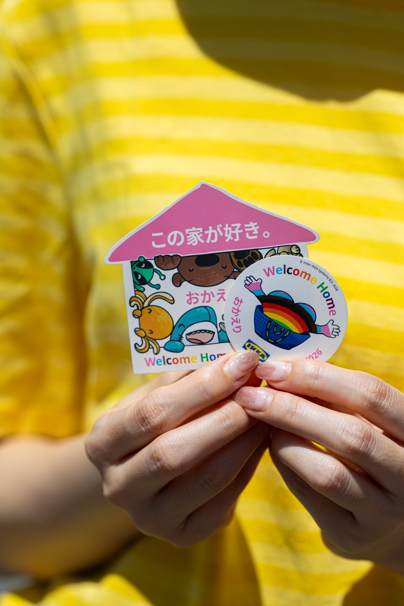 黄色いボーダーの服の前で両手が、Welcome Homeロゴとイケアの青いバッグとレインボーが描かれた家形カードと丸いバッジを持っている。