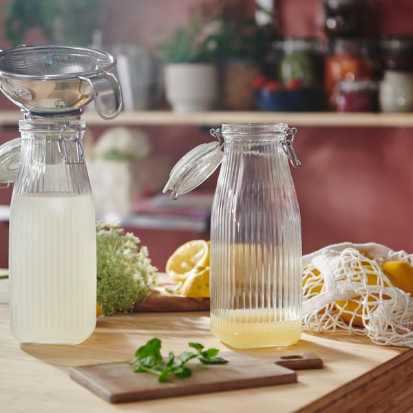 Two KÖSSEBÄR bottle shaped jars with lids, part-filled with yellowish liquid, sit on a worktop. One jar has a funnel in it. 
