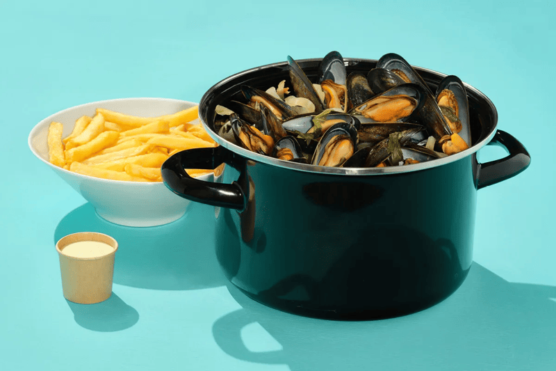 marmite de moule posée à côté d'une assiette de frite et d'un gobelet de mayonnaise, sur un fond turquoise
