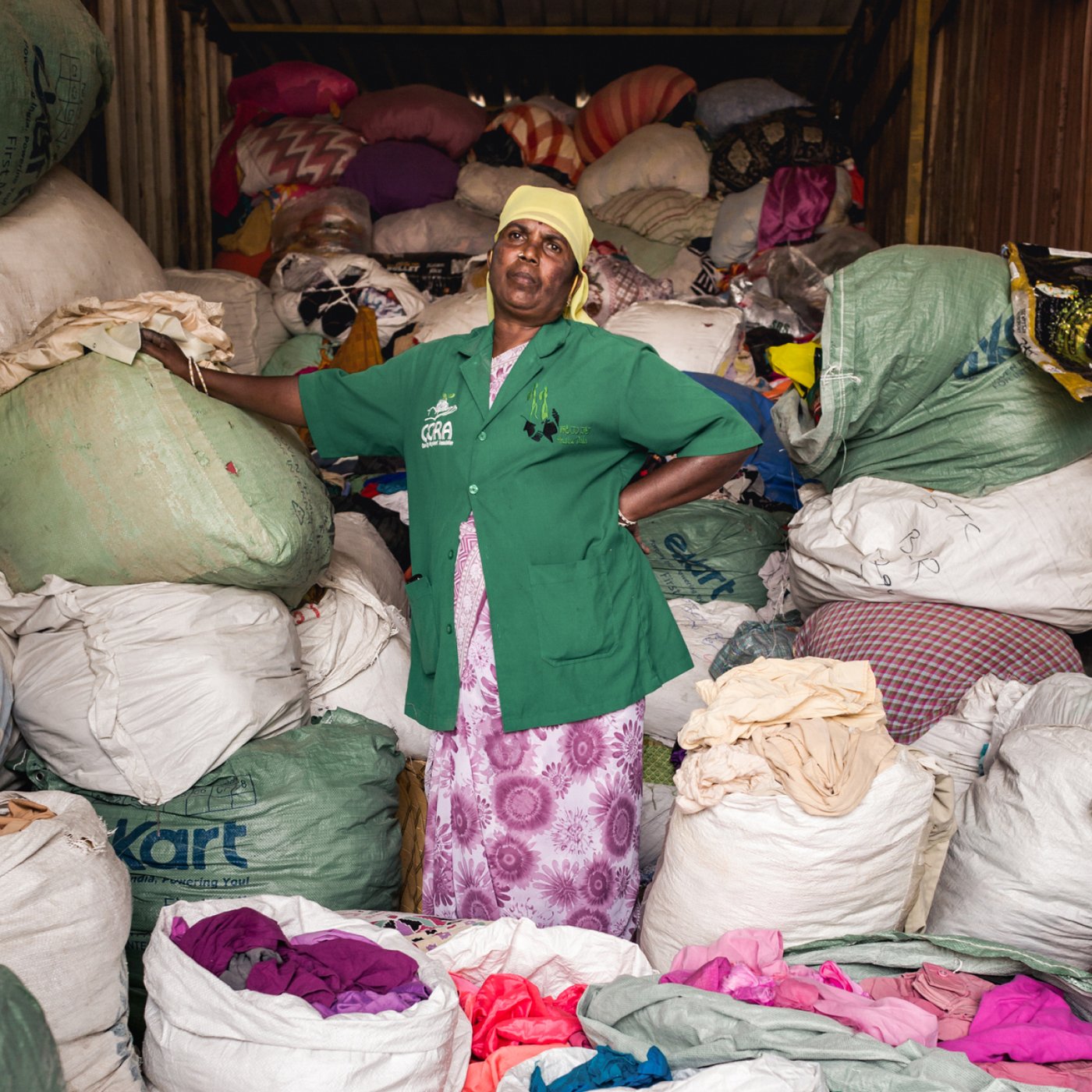 Osoba stoji u kontejneru napunjenim velikim vrećama i gomilama različitih tkanina. Osoba nosi zelenu košulju i šarenu suknju i izgleda kao da radi na sortiranju materijala.