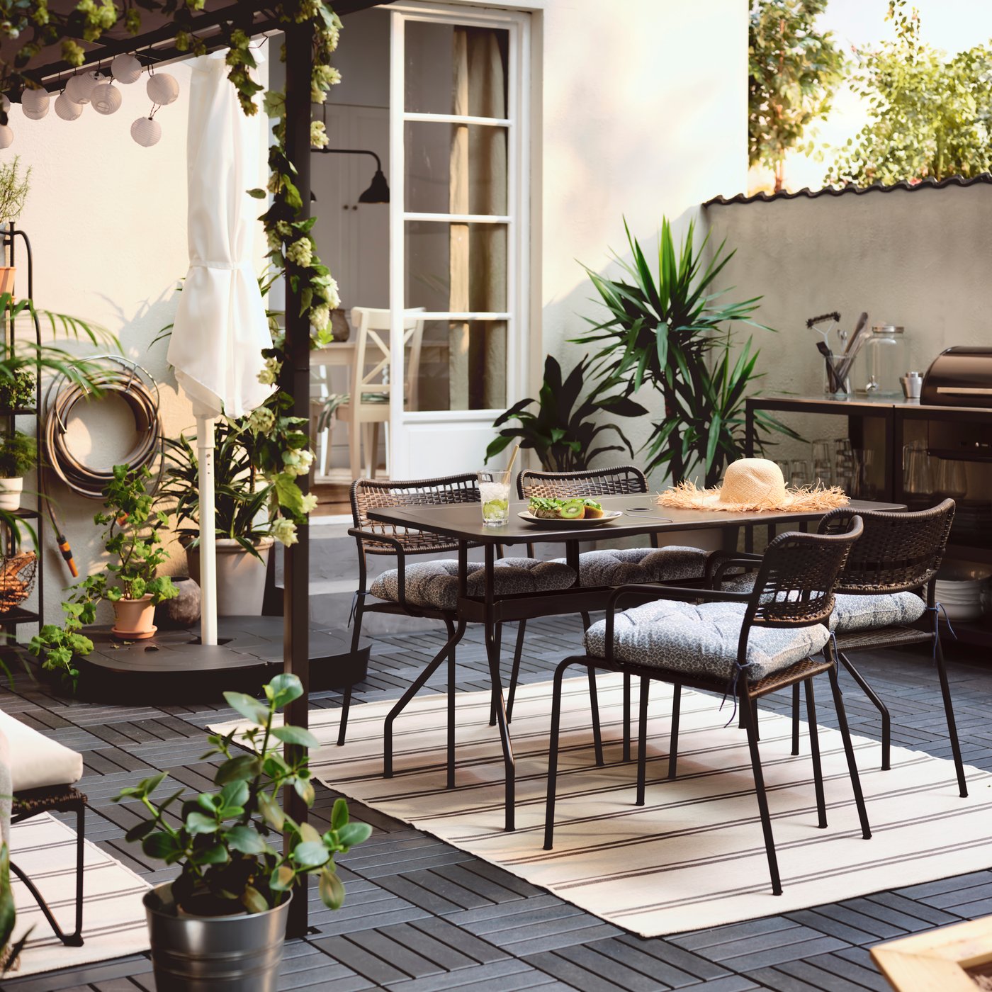 A terrace with a VIHOLMEN table surrounded by four LÄCKÖ chairs. An outdoor kitchen with charcoal barbecue is by the wall.