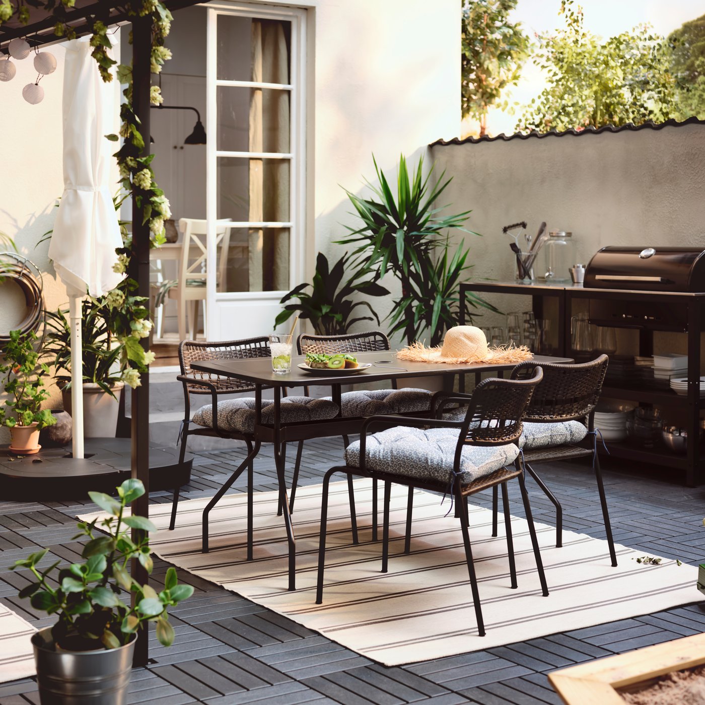 Una terrazza con un tavolo VIHOLMEN circondato da quattro sedie LÄCKÖ. Vicino alla parete, una cucina all'aperto con un barbecue a carbonella.