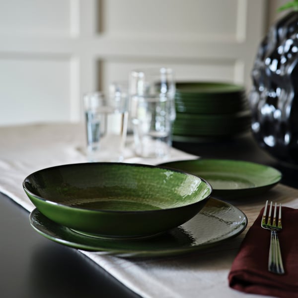 Une assiette creuse verte PELARKAKTUS est posée sur une table à côté de verres et d'assiettes empilées dans un coin repas stylé.
