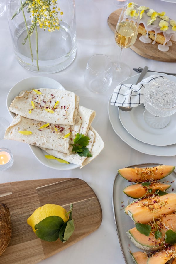 On a table are serving bowls and a board with sliced melon, wraps and other appetizers