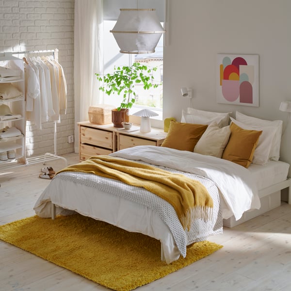 A bright bedroom with a white bed, yellow throw and yellow ÄRENDE rug sits beside wooden storage units against white walls.
