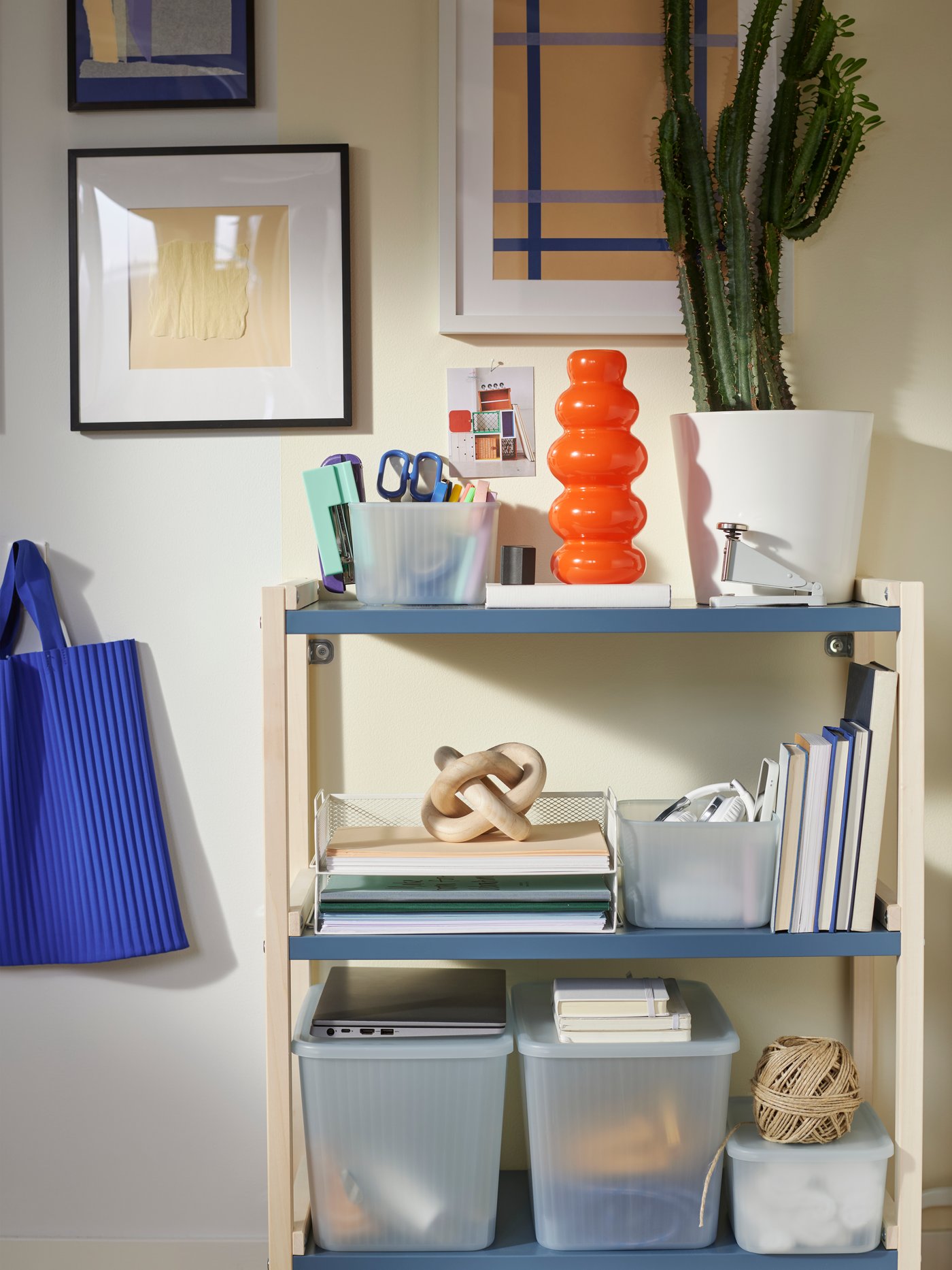 An open shelving unit with office supplies, storage boxes, a cactus, a white DRÖNJÖNS letter tray, and a curvy, orange vase.