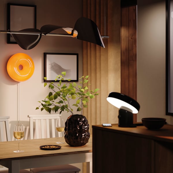 A black STOCKHOLM vase is standing on a table in a cosy dining area with several lamps, creating a soft atmosphere.