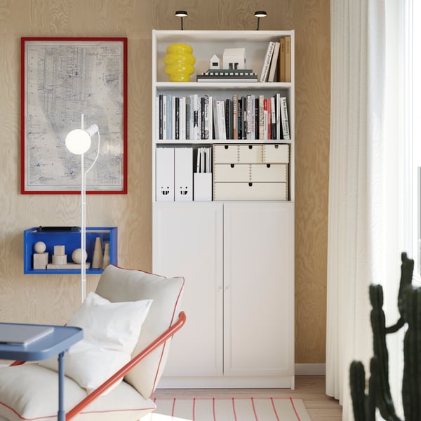A white BILLY bookcase and a small blue cabinet hanging next to it, with a white floor lamp standing in the front.