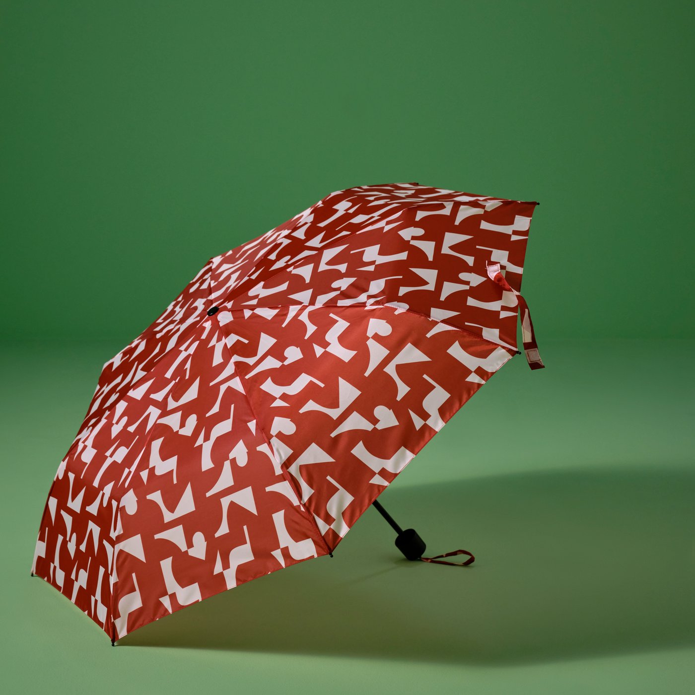 Rdeče-bel KNALLA zložljiv dežnik je odprt in leži na zeleni površini.