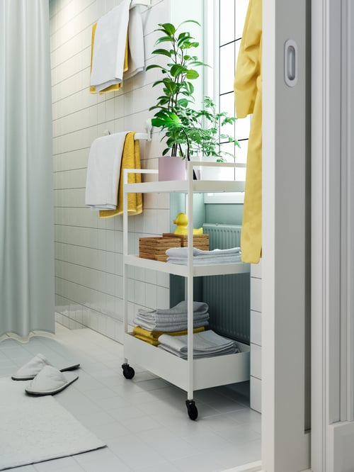 A white tiled bathroom. A NISSAFORS trolley with towels, a yellow rubber duck and a plant are placed on the shelves. 