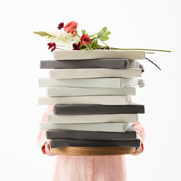 A person is holding a tall stack of JUSTINA chair pads in front of them. On top of the stack, there’s a bouquet of flowers.
