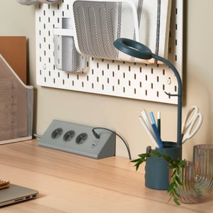 Birou IKEA ordonat, cu priză multiplă gri integrată pe blat, lampă de lucru, suport pentru creioane și panou perforat pentru organizare, ideal pentru un spațiu de lucru funcțional și sigur.