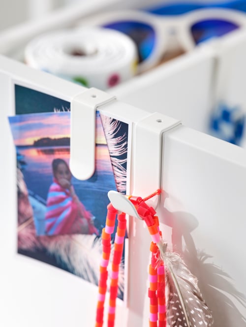 Detaliu al unității albe de depozitare cu cârlige albe versatile din oțel, cu cleme care conțin accesorii și fotografii.