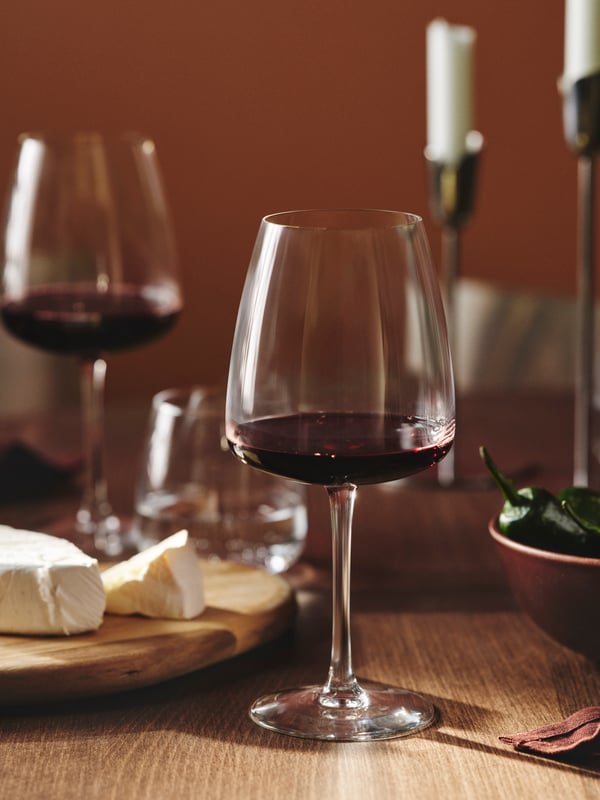 A DYRGRIP red wine glass, half filled, is standing on a wooden table next to a cheese platter and a bowl of green chillis.