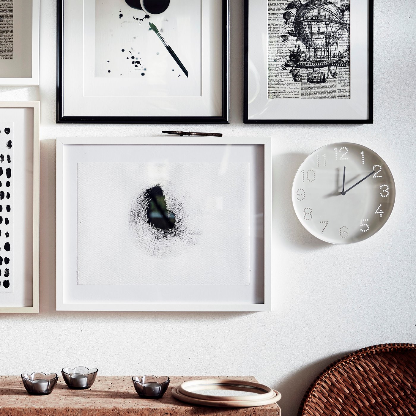 A light, softly sunlit wall surface with a white TROMMA wall clock hanging next to a neat arrangement of framed art.
