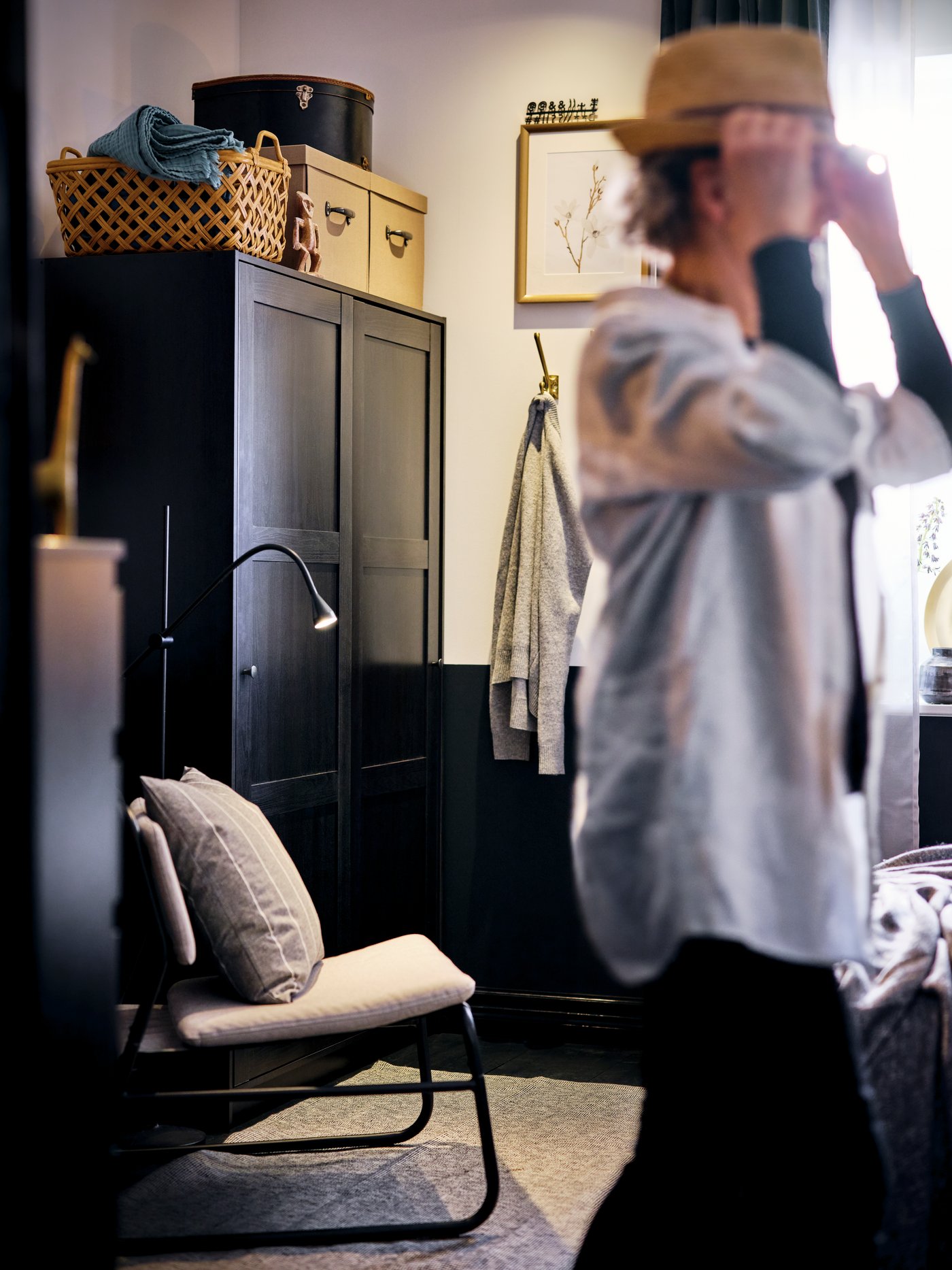 A child playfully dons a hat in a room furnished with a black-brown RAKKESTAD wardrobe and a light-grey LINNEBÄCK easy chair.