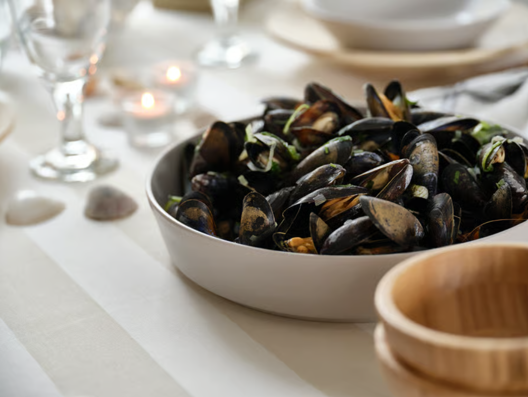 Schüssel mit gekochten Miesmuscheln auf hell gedecktem Tisch, umgeben von Gläsern und Geschirr.