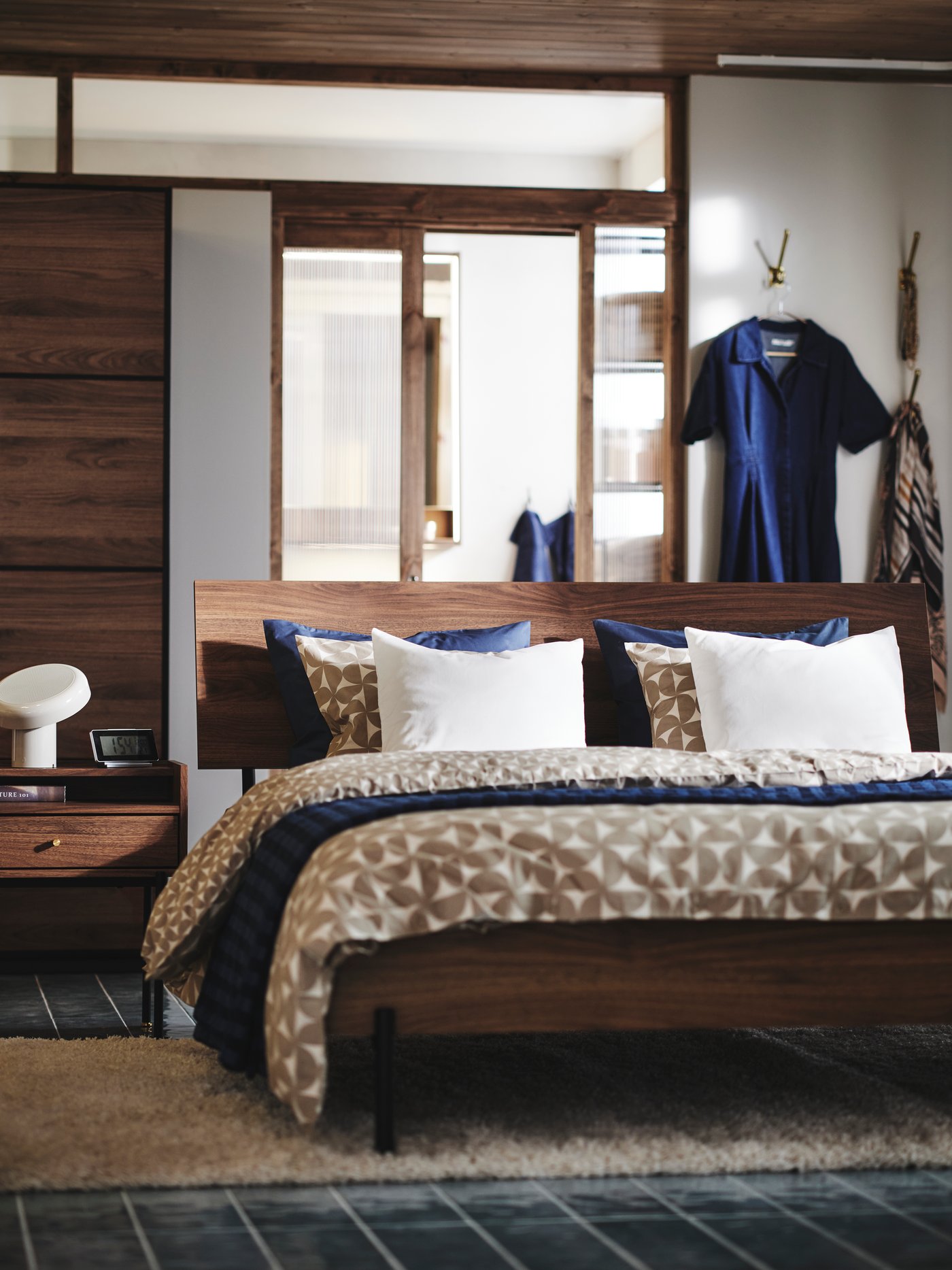 A brown walnut-effect RÅDMANSÖ bedside table and bed in a modern room with a PAX/MEHAMN wardrobe and wall hooks behind it.