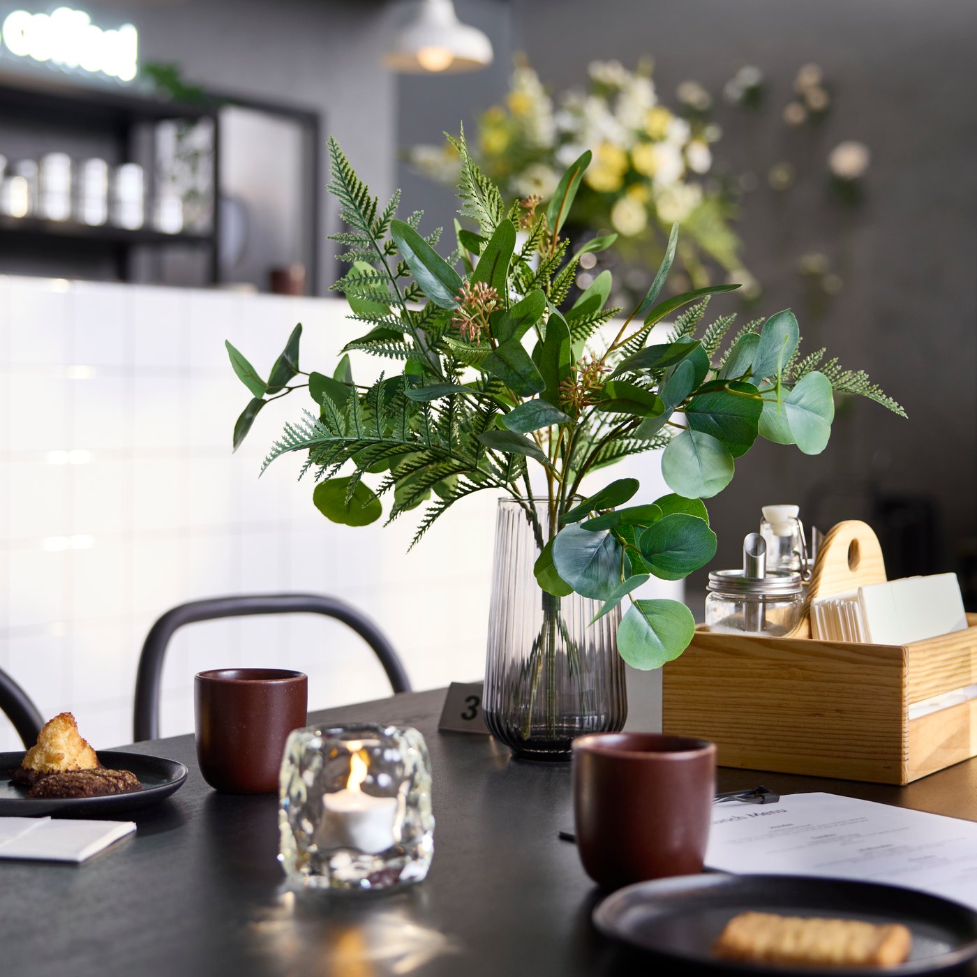 Gros plan sur une table noire avec un bouquet artificiel SMYCKA dans un vase en verre gris.