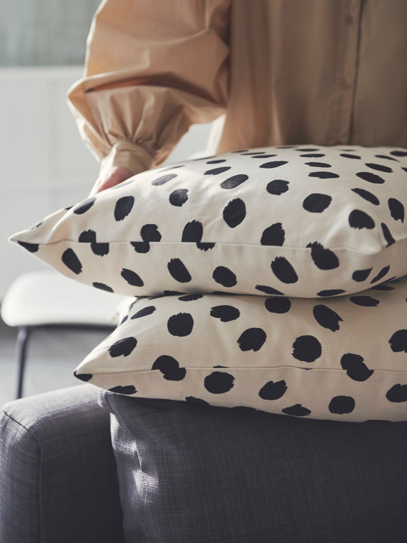 A person tends to two stacked cushions in ODDNY off-white-and-black dot-pattern cushion covers placed on a FRIHETEN sofa-bed.