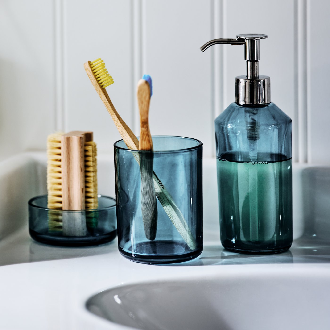 طقم حمام زجاجي من ثلاث قطع SKISSEN بجانب حوض الغسل، يحمل فرشاة أظافر وفرش أسنان وصابون سائل.