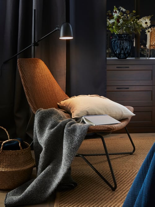 A scooped, dark brown/black RÅDVIKEN armchair that has a natural fibre seat and steel frame with a book, cushion and throw.