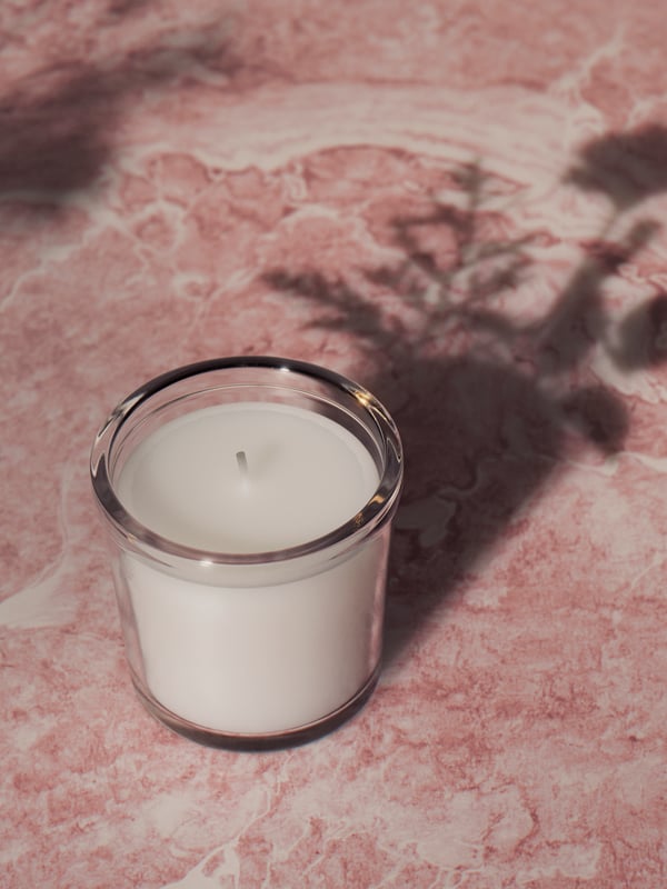 An ADLAD scented candle in a ceramic jar and a clear vase with leafy branches on a dining table close to a window.