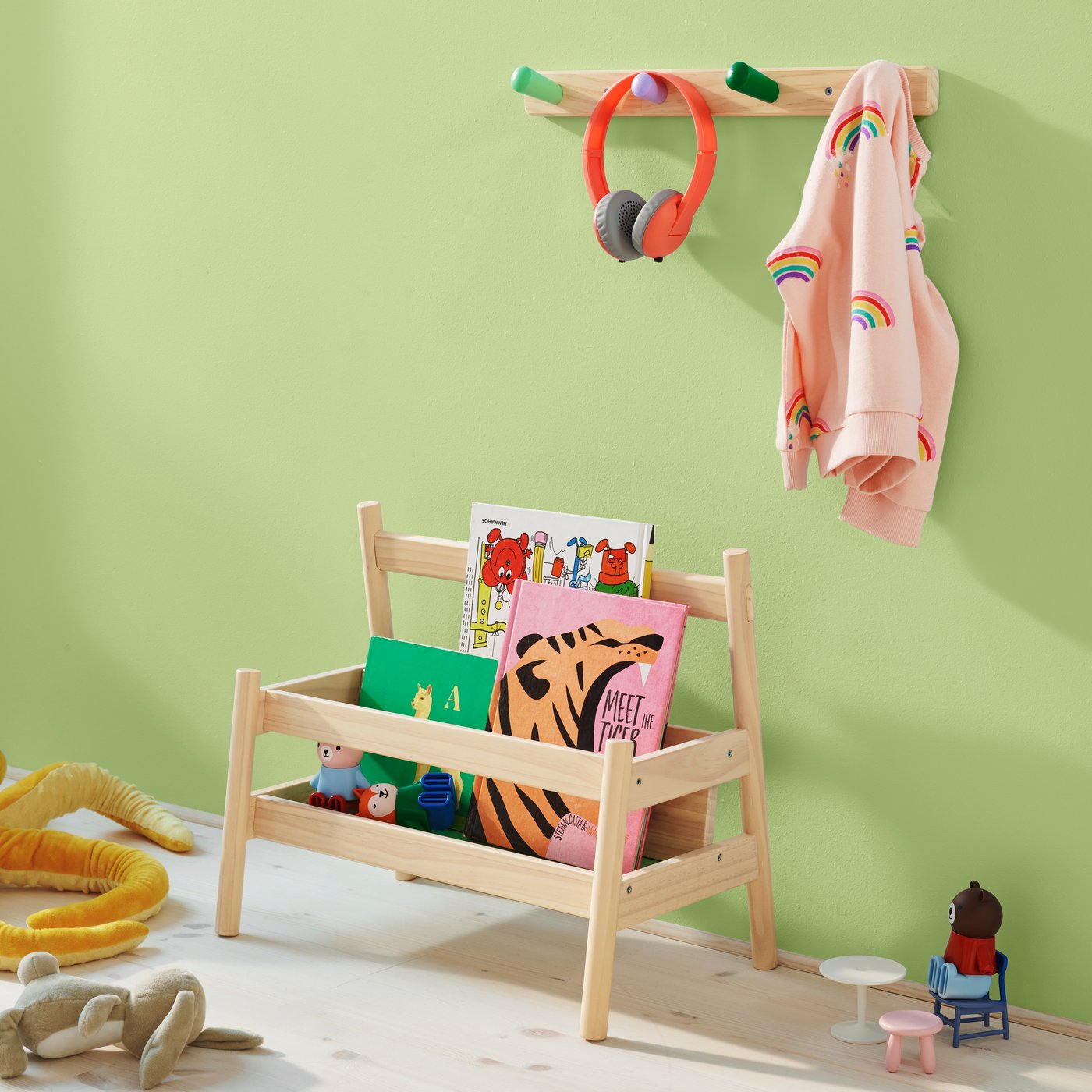 ライトグリーンの壁の子供部屋。木製の本棚にはカラフルな絵本やおもちゃが並び、壁のコートラックには虹柄のピンクのパーカーとヘッドフォンが掛けられています。床には遊び道具があり、明るく整理された空間です。