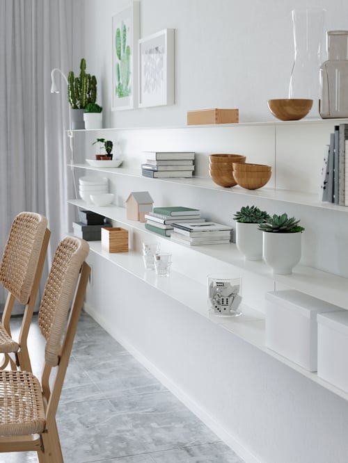 White wall-shelves with serving bowls, white plant pots, books and vases, set next to two beige chairs.