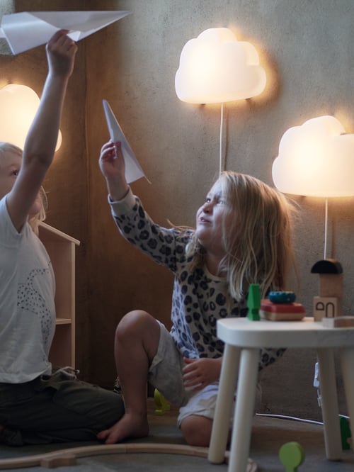 Two young girls playing with paper airplanes in front of wall lighting, beside a white wooden stool with toys on it.