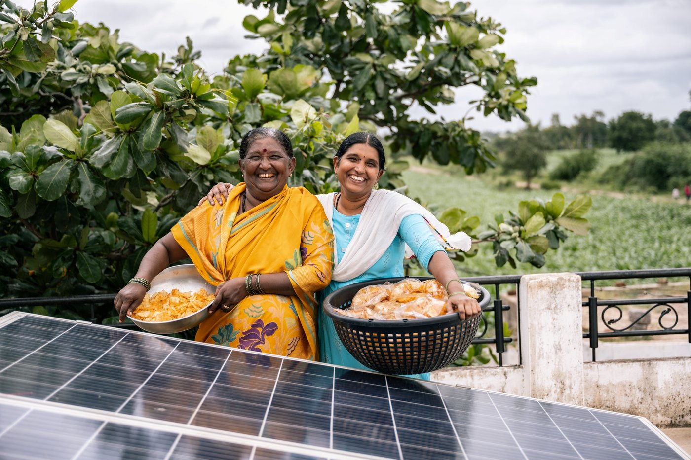 Two people stand next to solar panels and hold up baskets of vegetables against a lush green background.