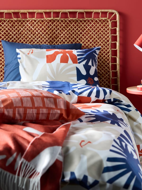 Een slaapkamer met een rotan bedframe en gekleurd beddengoed in rood-wit-blauw met een bloemenpatroon erop en een beige TIDTABELL vloerkleed.