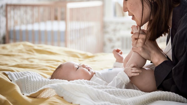 A baby lying on a bed with a white blanket, while an adult is attending to them; the background shows a crib in a well-lit room.