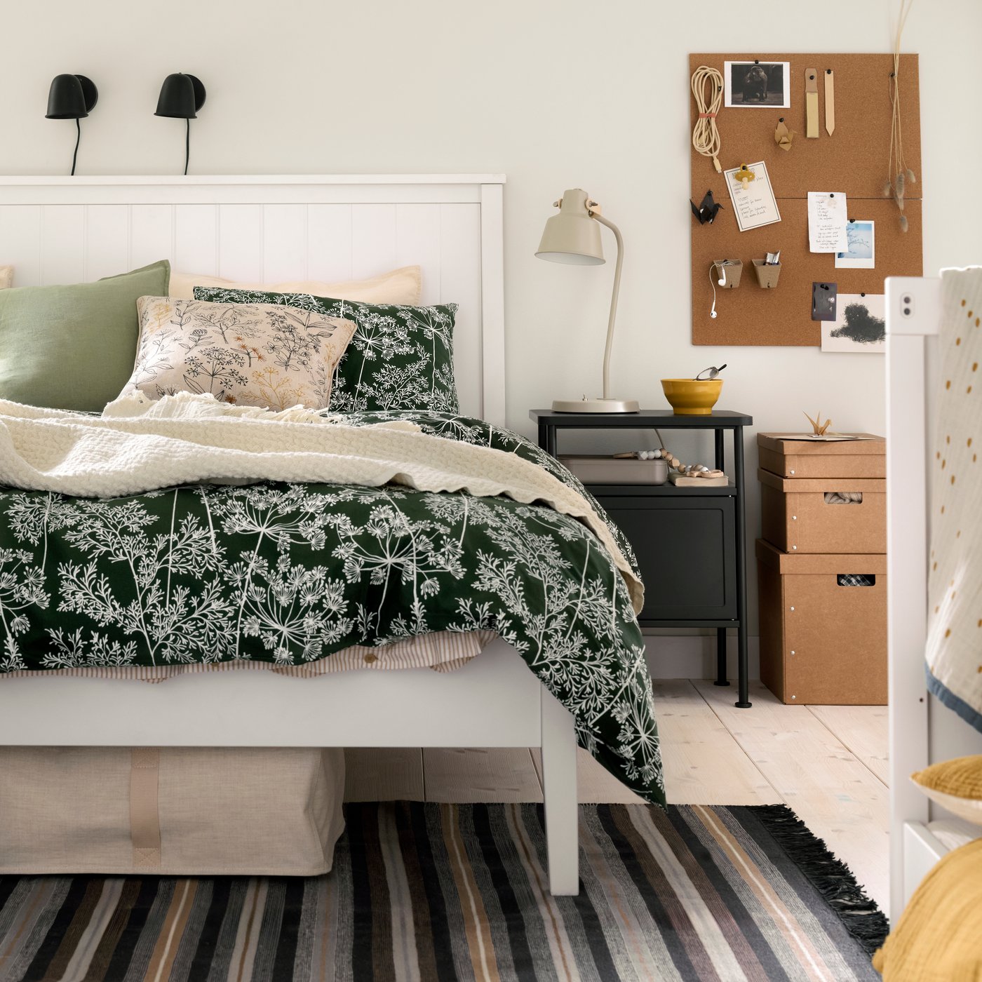 Een slaapkamer met een houten GULLABERG bed met een dekbedovertrek met bloemenprint, een donkergrijs nachtkastje en de rand van een babybed.